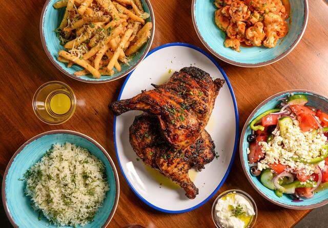 A top-down view of a spread of Greek cuisine on a wooden table. The center features a white platter with two large pieces of charred, herb-rubbed roasted chicken. Surrounding the chicken are several turquoise bowls containing seasoned french fries topped with herbs and cheese, a portion of white rice mixed with fresh dill, buffalo-style cauliflower florets, and a large Greek salad with tomatoes, cucumbers, olives, and a generous topping of crumbled feta cheese. Two small side containers of white sauce (tzatziki) and oil are also visible.